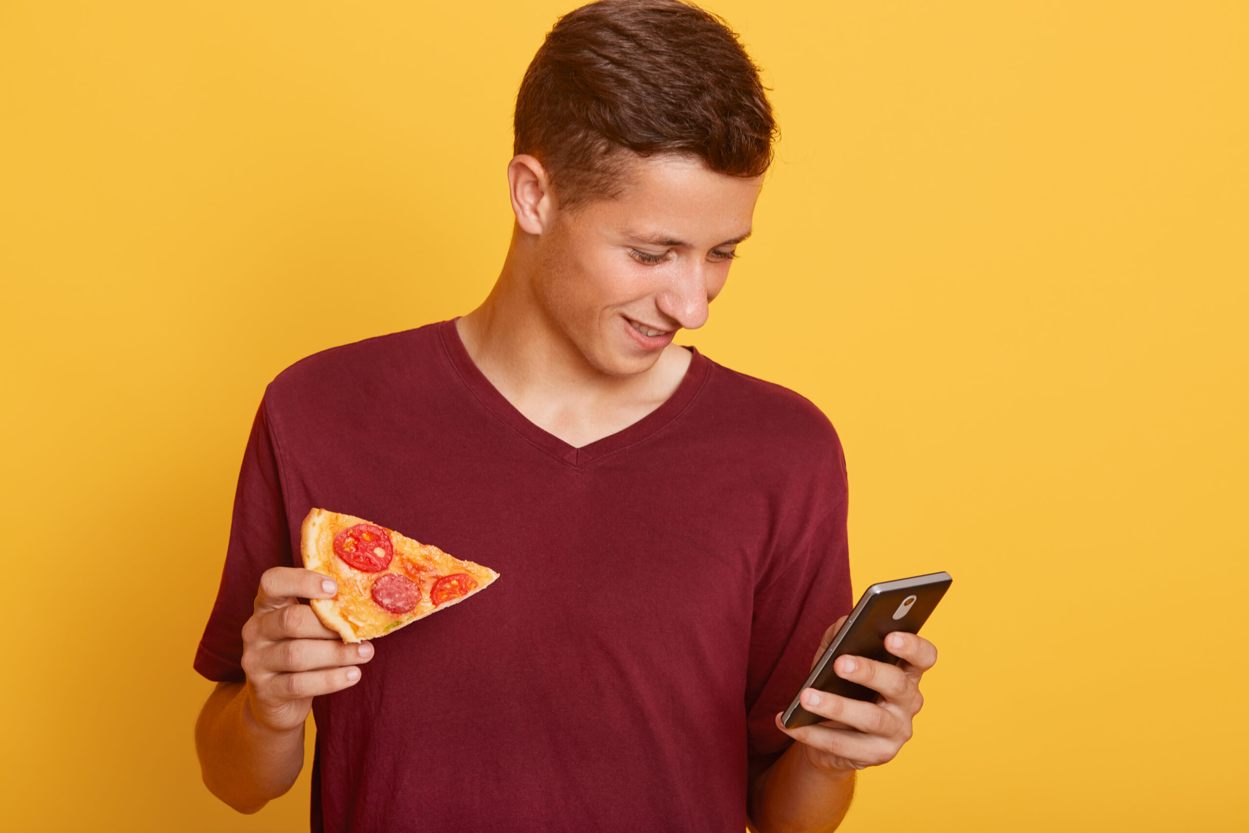 Oferte Pizza Iza photo of busy curious joyful teenager holding his smartphone, using his device, checking social networking sites, having piece of pizza in one hand, wearing red t shirt. youth and free time concept.